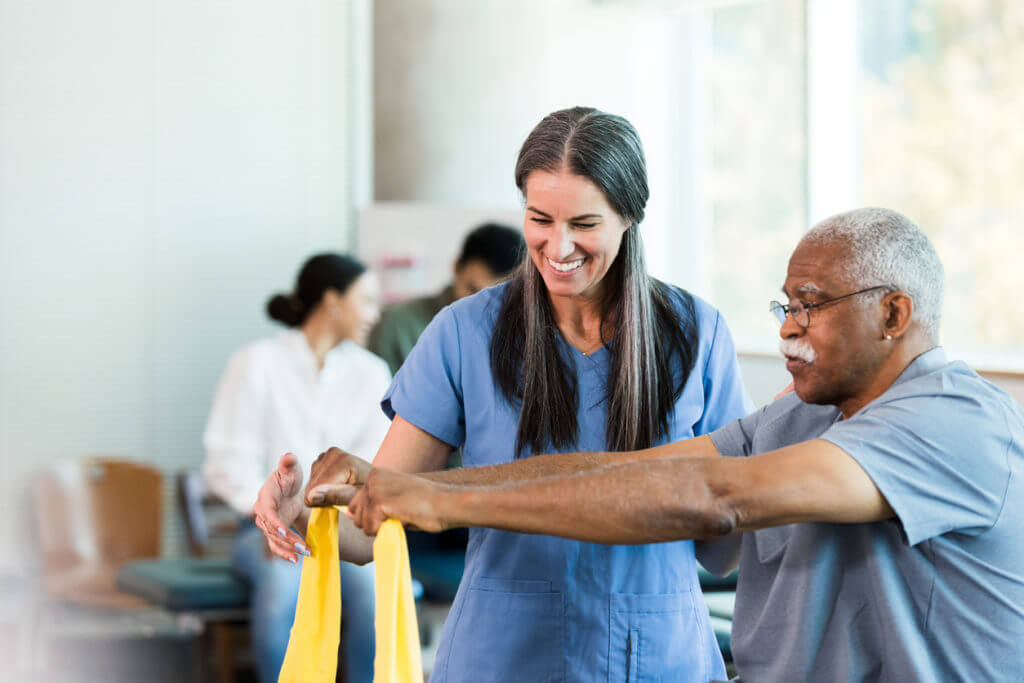 Therapist smiles as senior man learns to use elastic band