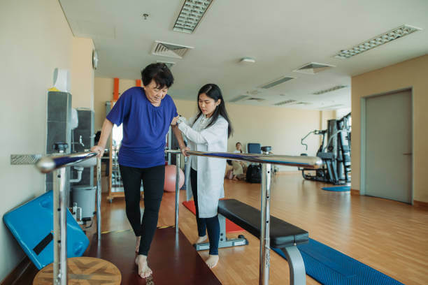 Asian chinese senior women practicing, holding parallel bars and walking while physical therapists helping at side in a rehab centre at hospital