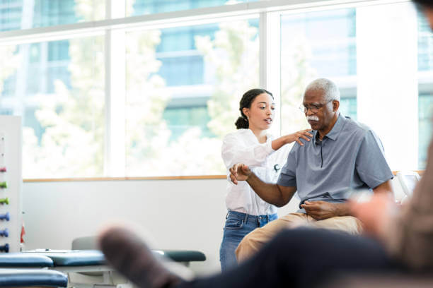 As the senior man grimaces, the young adult female physical therapist manipulates his injured shoulder.