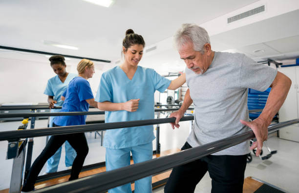 Diverse team of physiotherapist helping senior patients walk between parallel bars all looking happy and smiling