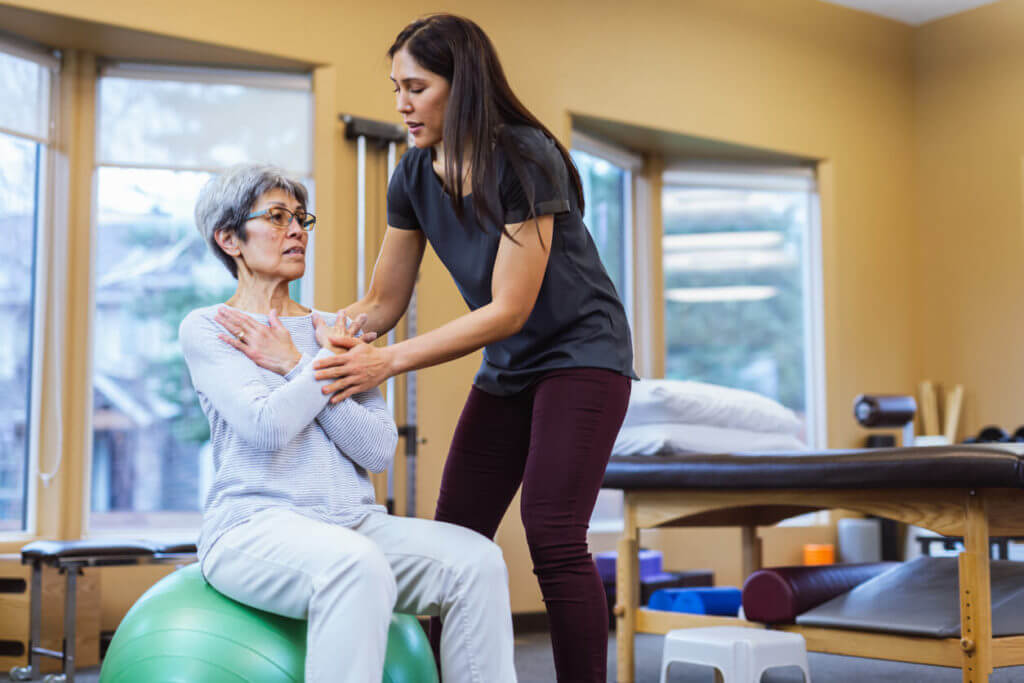 During a physical therapy session, a senior patient sits on a yoga ball with the assistance of a physical therapist.