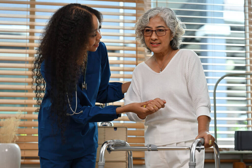 An African female physical therapist helps and supports an elderly stroke patient woman standing with a walker at home.