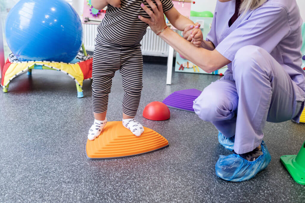 A child is balancing on a sensory board during a pediatric physical therapy session.