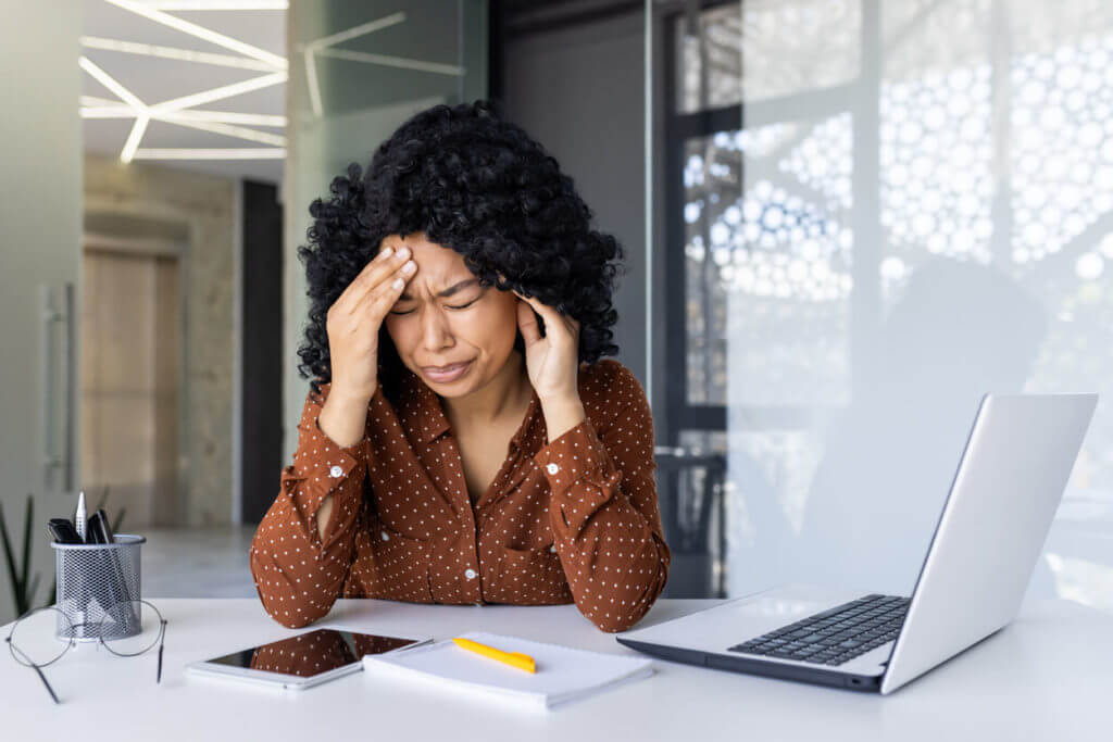 A stressed young woman is experiencing a headache at her work desk due to a migraine.