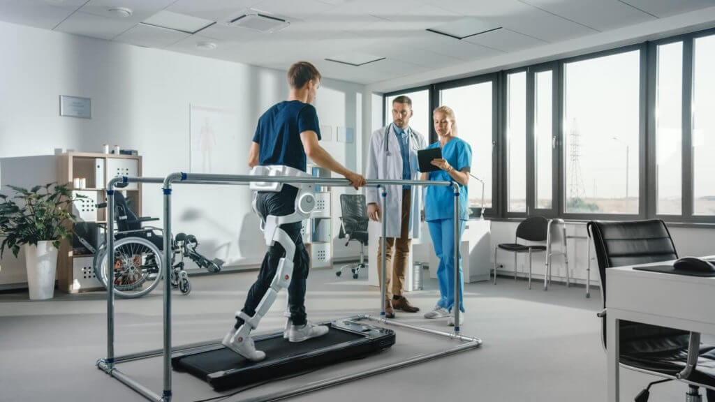 A patient with an injury walks on a treadmill wearing an advanced robotic exoskeleton in a modern hospital's physical therapy department.